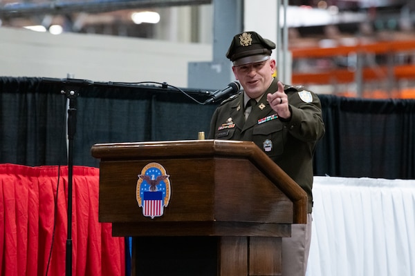 A man wearing a military uniform stands on stage behind a podium speaking into a microphone