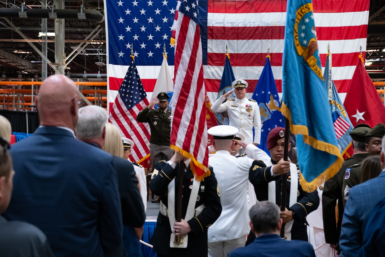 Three men wearing military uniforms salute flags while standing on a stage.