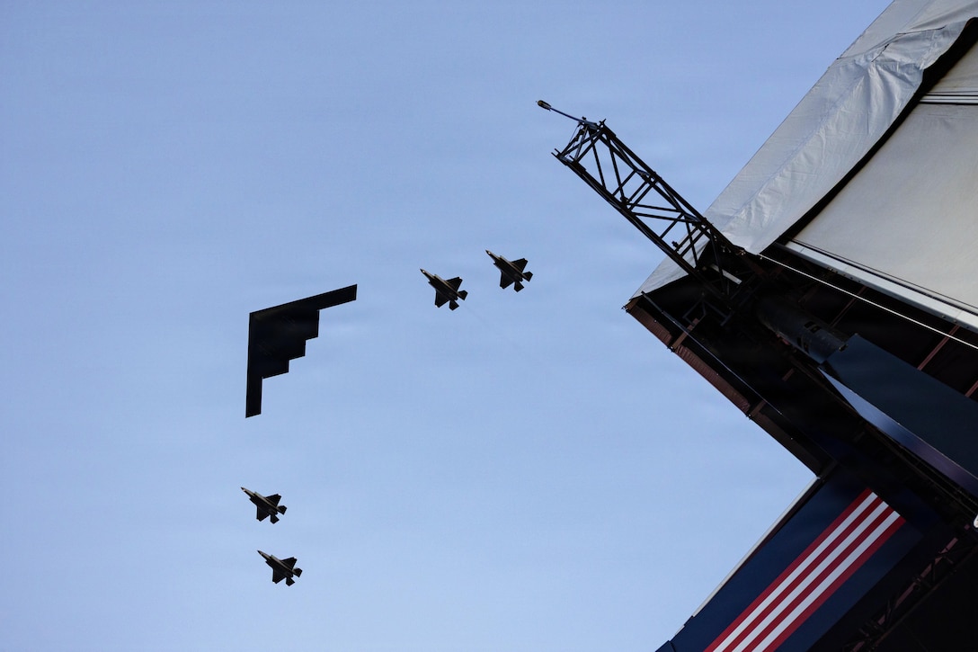 A stealth bomber and four fighter jets are in silhouette and in formation in the air. A stage is below them nearby.
