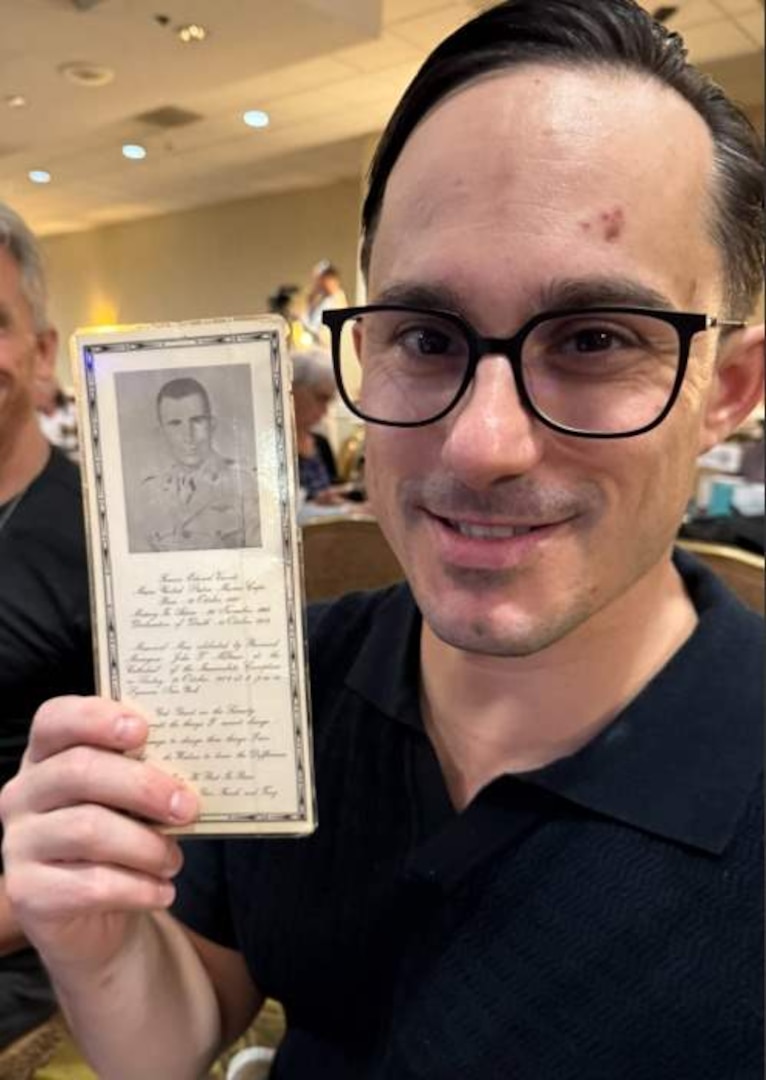 A man in casual attire holds up a photo of another man while seated in a ballroom.