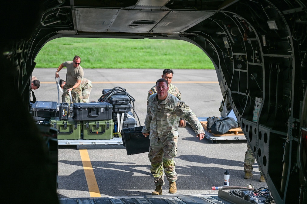 Two airmen approach the opening of a grounded helicopter while carrying a container. Each airman has one hand on the container.