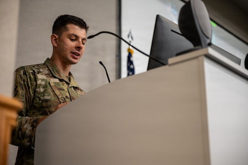A man in a U.S. Air Force camouflage uniform stands at a white lectern and speaks into a microphone.