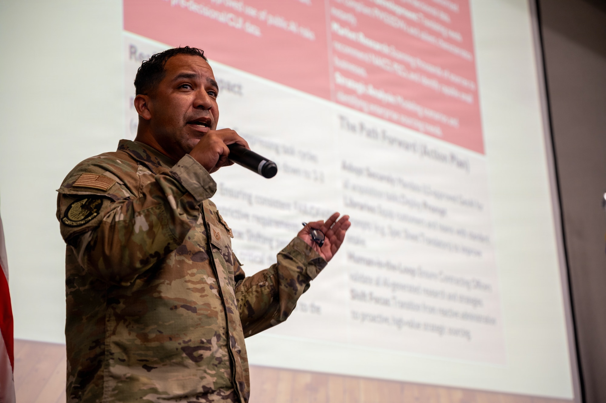 A U.S. Airman in a camouflage uniform gestures while speaking into a handheld microphone, with a presentation slide visible behind him.
