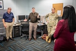 Four people are gathered in an office space. An Army officer in a brown jacket is speaking to the group, which includes another man in a polo shirt, a woman in a military uniform, and a woman in a red blazer who is seen from the back.