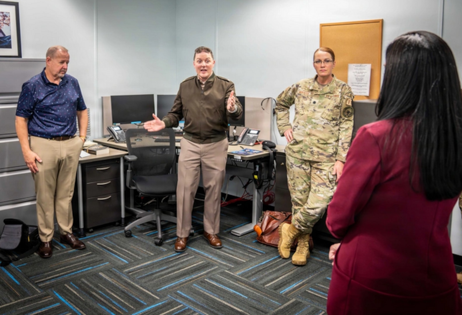 Four people are gathered in an office space. An Army officer in a brown jacket is speaking to the group, which includes another man in a polo shirt, a woman in a military uniform, and a woman in a red blazer who is seen from the back.