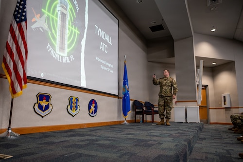A U.S. Airman in a camouflage uniform stands on a stage, using a remote to present a slide about "TYNDALL ATC." An American flag and Air Force emblems are also on stage.