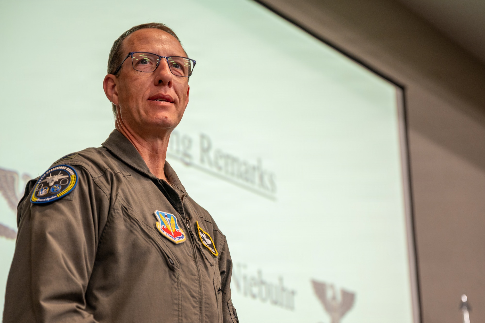A close-up photo of a U.S. Air Force pilot in a flight suit and glasses, standing in front of a presentation screen.