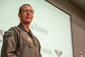 A close-up photo of a U.S. Air Force pilot in a flight suit and glasses, standing in front of a presentation screen.