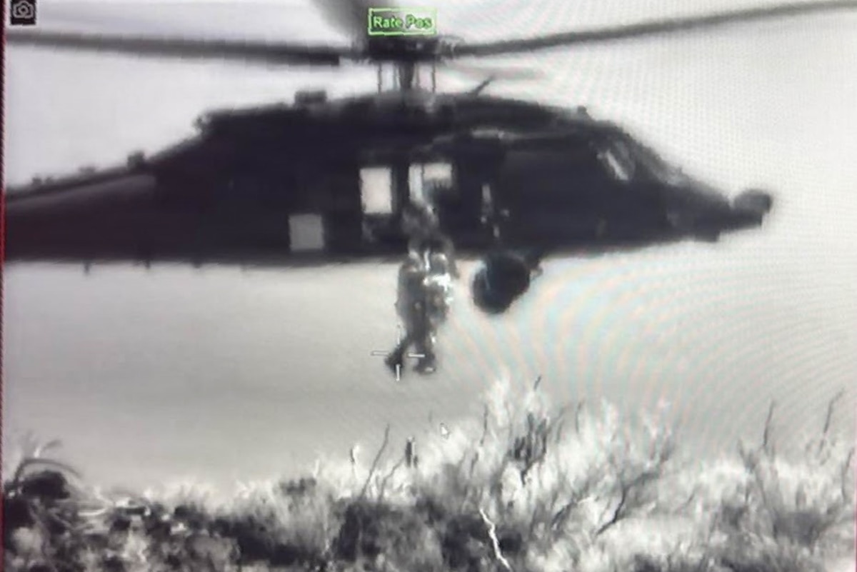 A grainy, black-and-white image of a helicopter hovering over water with two people dangling from it and a third person assisting as they are hoisted into the aircraft.