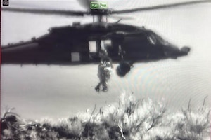 A grainy, black-and-white image of a helicopter hovering over water with two people dangling from it and a third person assisting as they are hoisted into the aircraft.