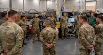 A group of uniformed U.S. Air Force personnel stand in a circle inside a maintenance facility surrounded by equipment and tires.