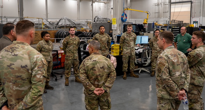 A group of uniformed U.S. Air Force personnel stand in a circle inside a maintenance facility surrounded by equipment and tires.
