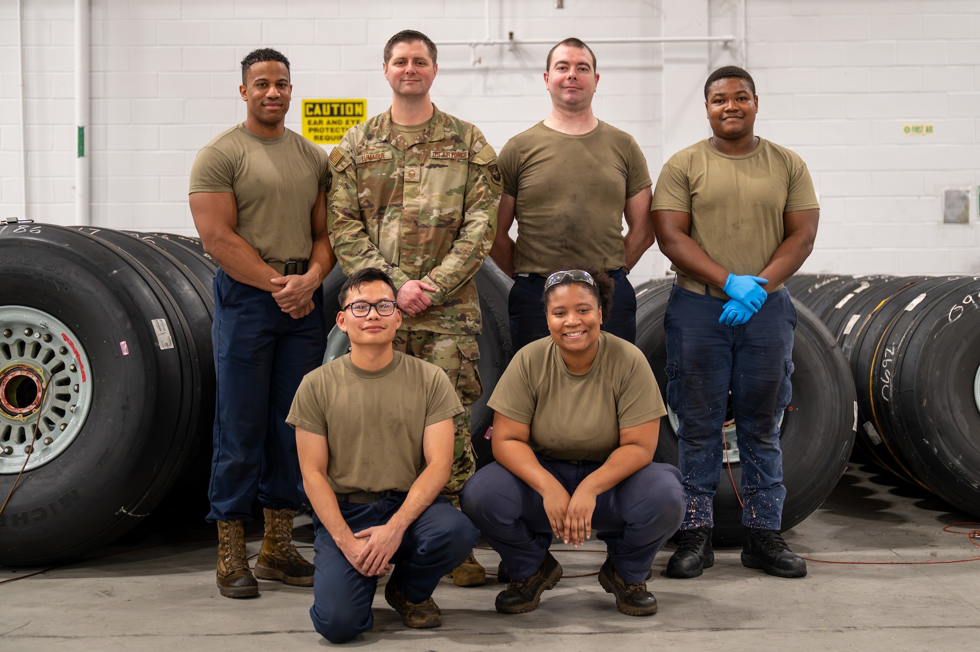 Seven U.S. Air Force personnel pose for a group photo, standing and kneeling, in the wheel and tire maintenance facility, in front of C-17 Globemaster III tires.