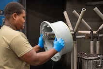 A U.S. Air Force maintainer wearing blue protective gloves removes a C-17 Globemaster III wheel rim from a rim washer in a maintenance shop.