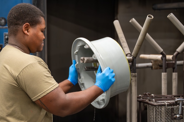A U.S. Air Force maintainer wearing blue protective gloves removes a C-17 Globemaster III wheel rim from a rim washer in a maintenance shop.