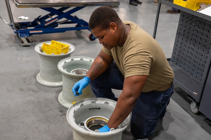 A U.S. Air Force maintainer wearing blue protective gloves kneels on the shop floor while assembling C-17 Globemaster III wheel rim components in a maintenance shop.