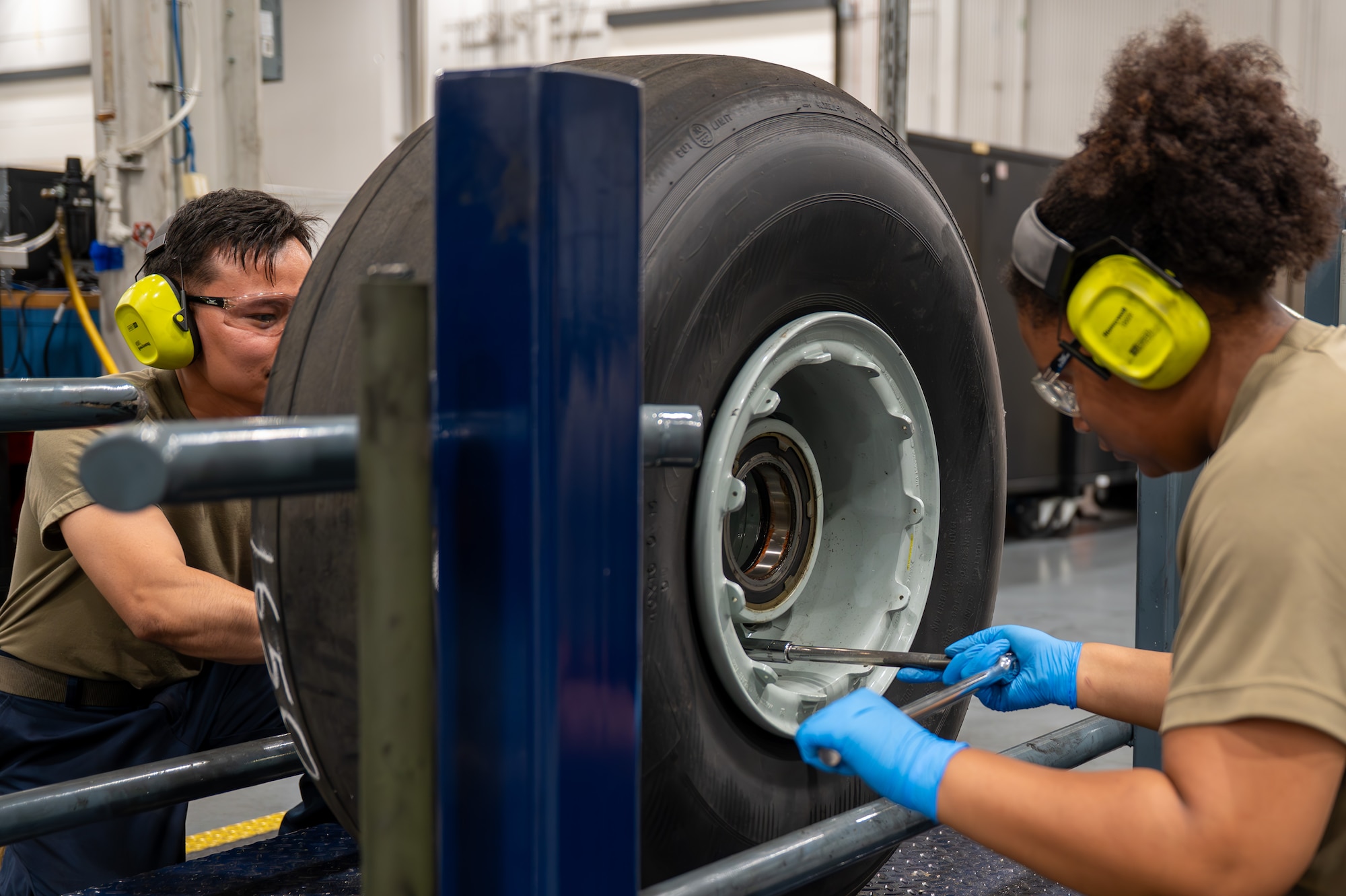 Two U.S. Air Force maintainers wearing hearing protection, safety glasses, and gloves work together to secure a C-17 Globemaster III tire using hand tools in a maintenance shop.