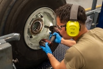 A U.S. Air Force maintainer wearing hearing protection, safety glasses, and blue protective gloves uses hand tools to tighten fasteners on a C-17 Globemaster III wheel during maintenance.
