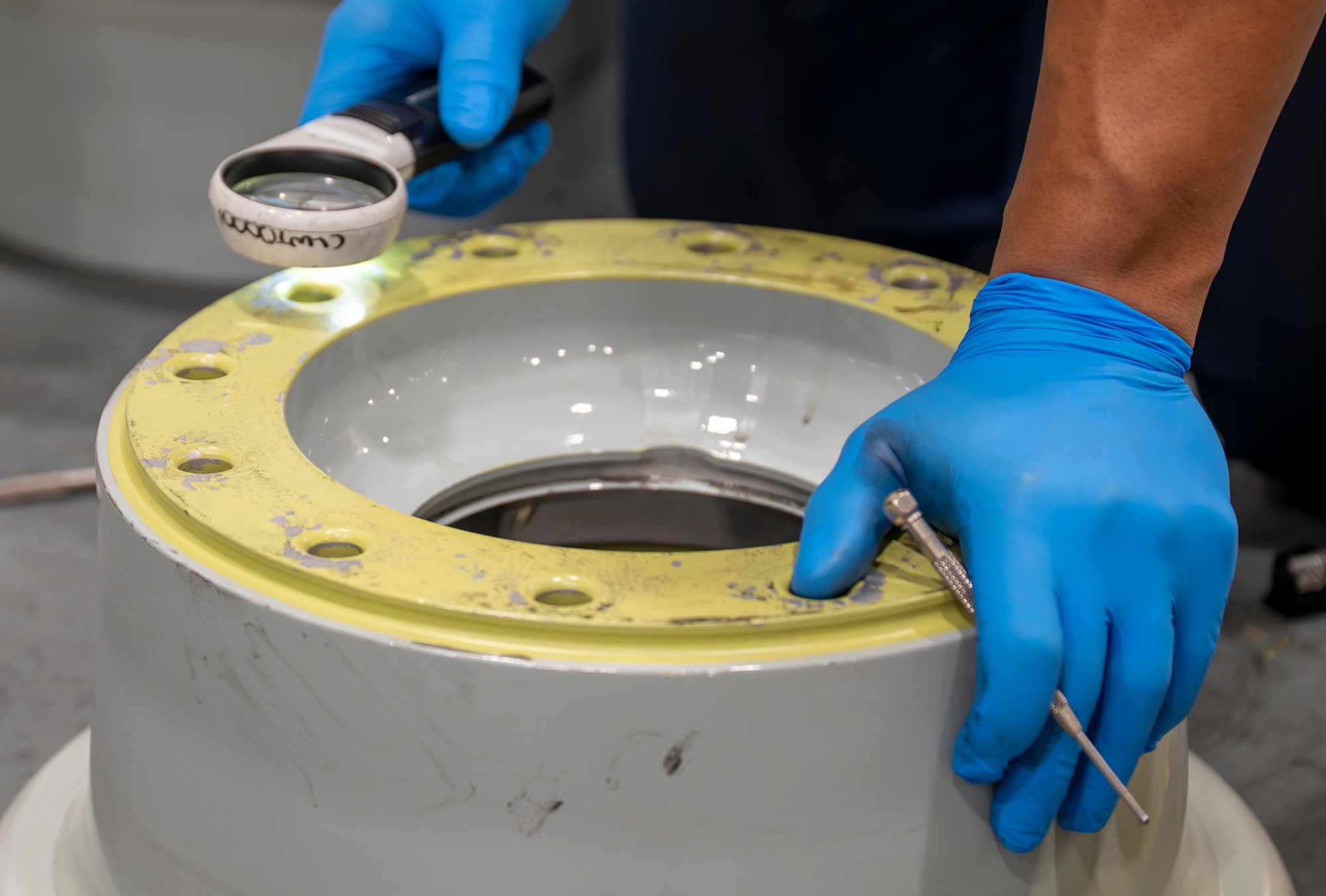 A technician wearing blue protective gloves inspects an C-17 Globemaster III wheel rim with a magnifying glass.