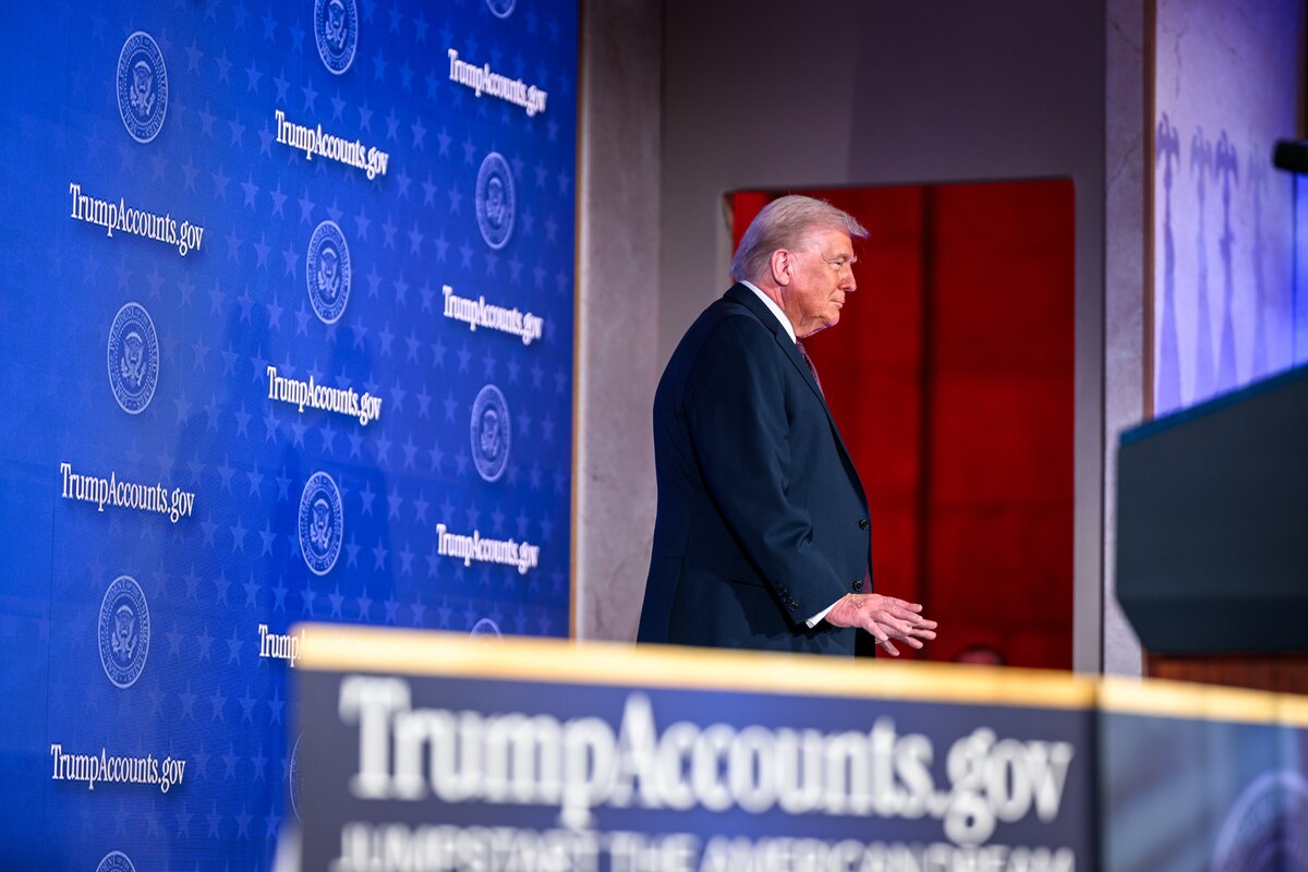 President Donald J. Trump stands on a stage in front of a blue wall adorned with TrumpAccounts.gov logos.