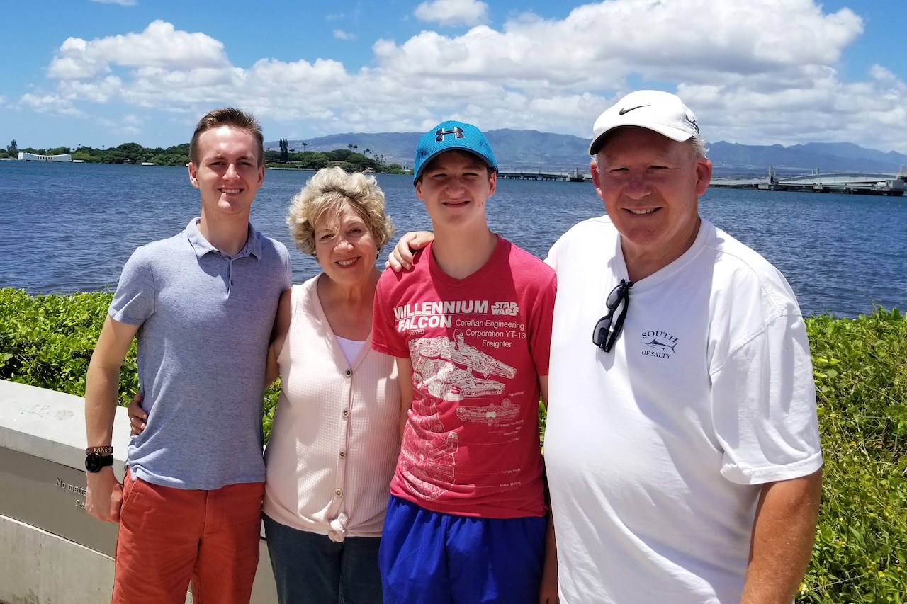 Four people in civilian attire pose for a photo outdoors during daytime, with a body of water and mountain range in the background.