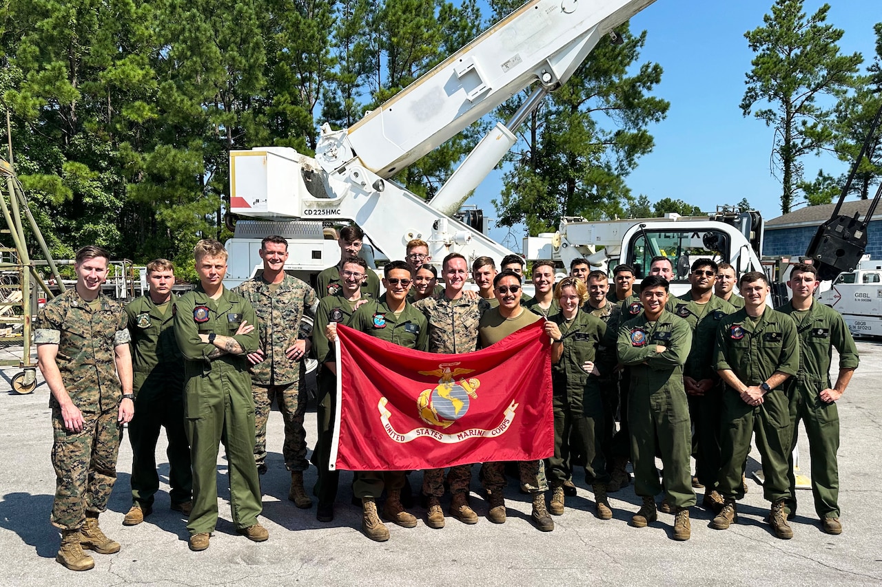 Marines stand as a group outdoors during daytime and smile for the camera, with two people in front holding a red Marine Corps flag.