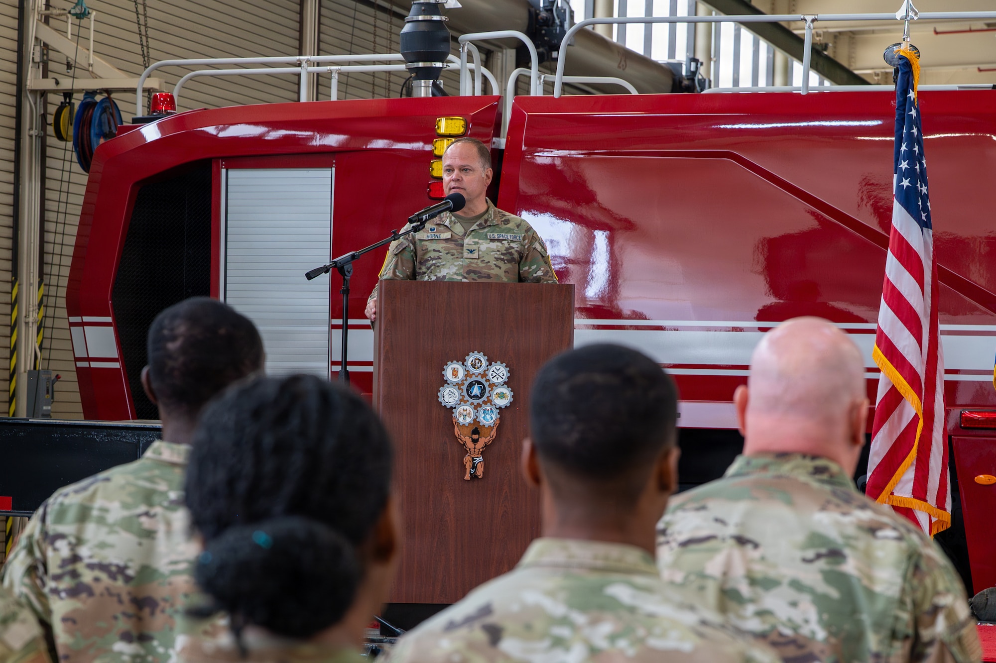 A person stands behind a podium and speaks while people stand in the foreground and listen