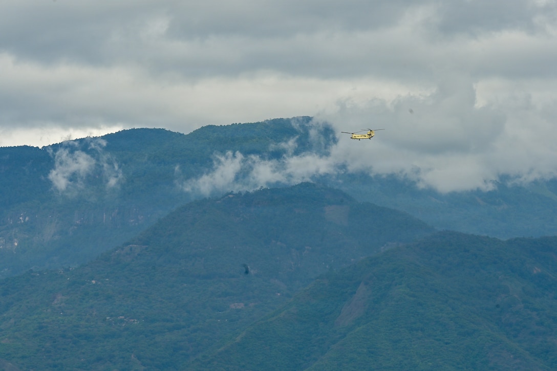 A military helicopter flies in the distance with mountains in the backdrop.