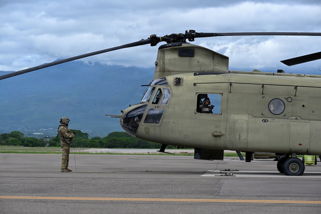A helicopter crew member stands in front of a helicopter on the flight line.