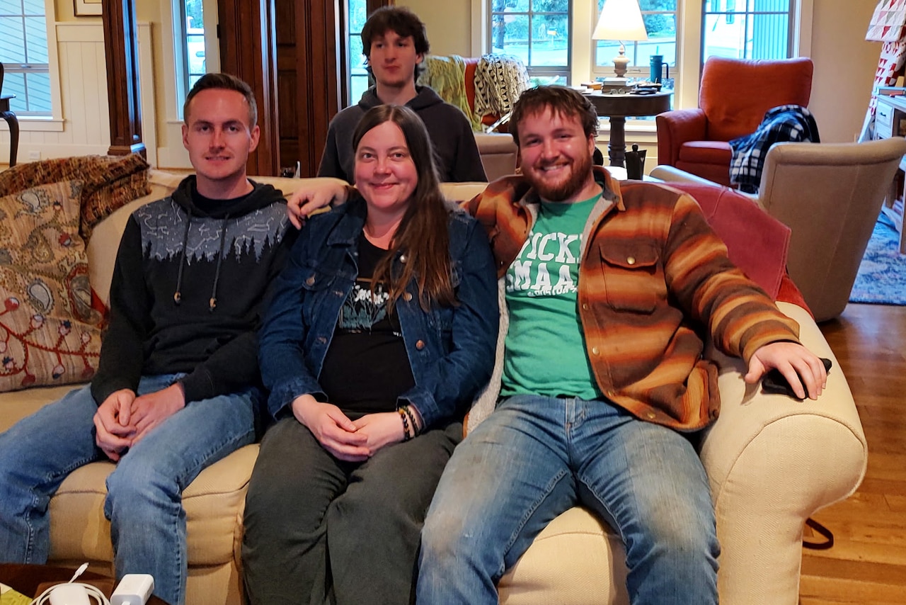 Three people in civilian attire sit on a couch, with another person in similar attire situated behind the couch, in a living room.