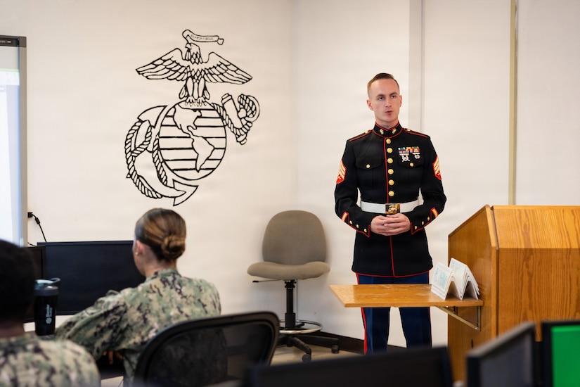 A Marine in a formal uniform stands in a classroom setting and speaks to people sitting at desks.