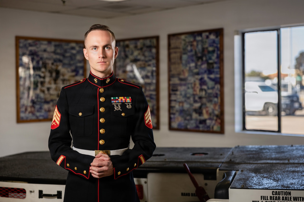 A Marine stands in a room indoors, with framed photos on the wall behind him and a window.