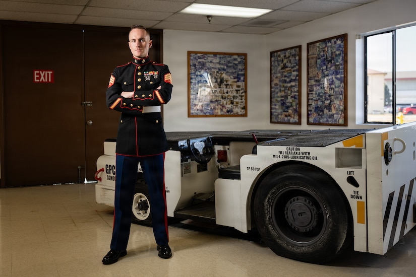 A Marine wearing a formal uniform stands with his arms crossed in a room near a short vehicle, with a window and framed pictures on the wall in the background.