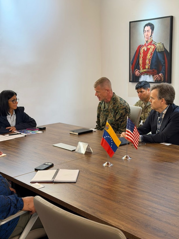 A man in a military uniform speaks to a woman in a dark jacket during a meeting at a table.