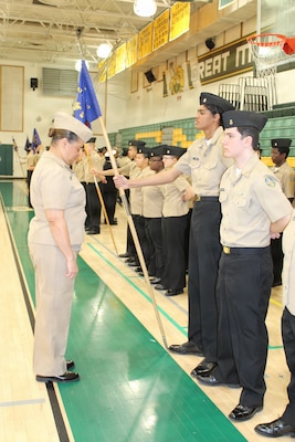 Cmdr. Nichole Benson, the Review Officer for the Great Mills High School Navy Junior Reserve Officers Training Corps (NJROTC) Annual Military Inspection, inspects the platoon guidon presented by her son. Benson, a nurse practitioner assigned to Naval Health Clinic Patuxent River, served as the guest of honor for the inspection, evaluating cadets on military bearing, uniform appearance, and general knowledge. The moment highlights the deep community and family ties between the local naval command and the school's NJROTC program.