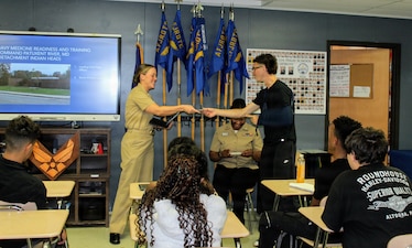 An Air Force Junior Reserve Officers Training Corps (AFJROTC) cadet presents a token of appreciation to Chief Petty Officer Amber Ronquille, assigned to Navy Medicine Readiness and Training Command (NMRTC) Patuxent River, at Chopticon High School. Ronquille, along with Hospital Corpsman 2nd Class Ifeoluwapo Olayemi (seated), had just finished presenting to the cadets on leadership and professional development as part of the command's spring community outreach initiative.
