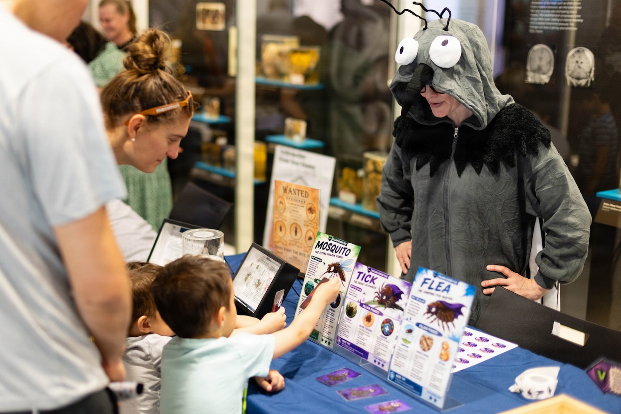 A woman in a bug costume talks to two kids as they look at displays on bugs.