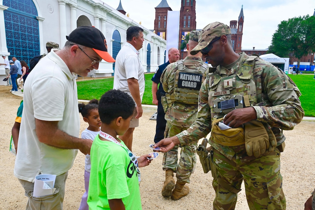 A soldier gives stickers to children and their parents near tall buildings during the day.