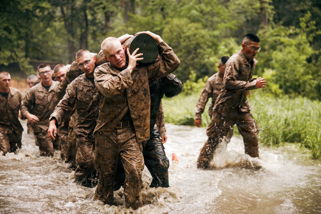 Marines walk through waters while carrying a log on their shoulders in a wooded area.