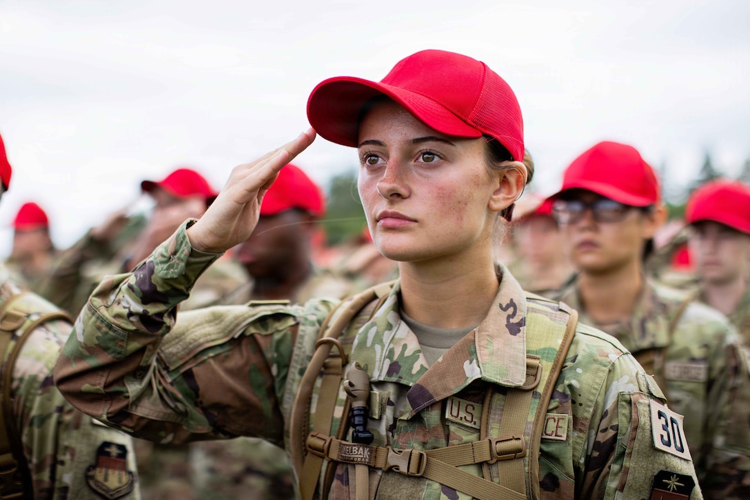 An Air Force basic cadet in a red hat salutes against a blurred background of dozens of other cadets in red hats.