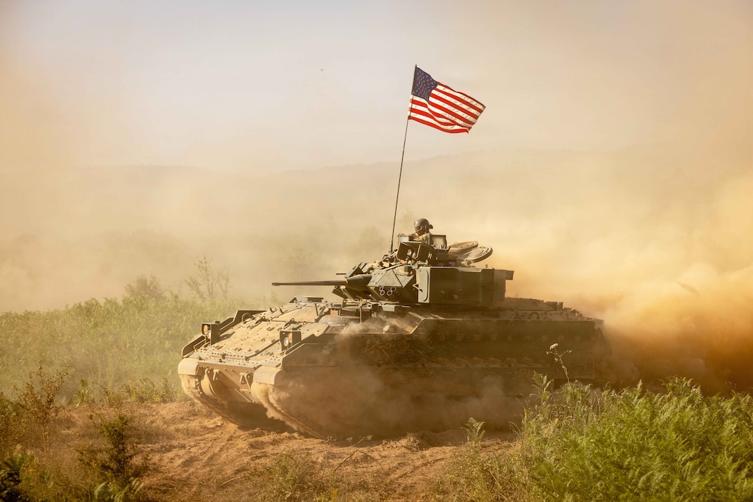 Dust fills the air as a tank drives through a desert displaying an American flag.