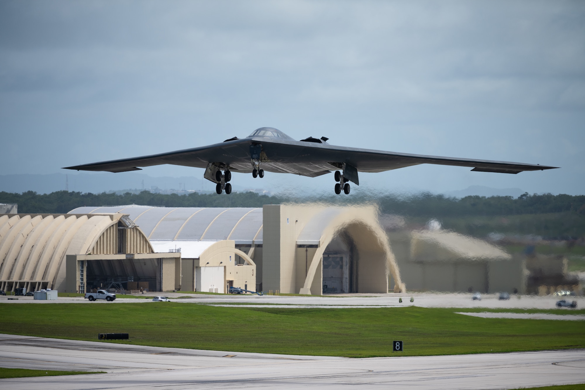 A stealth bomber takes off from an airfield with aircraft hangars behind it