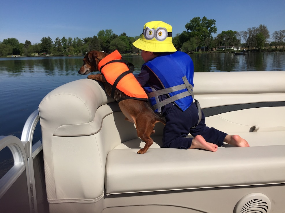 Child and dog, both wearing PFDs, on a pontoon, look over the water.