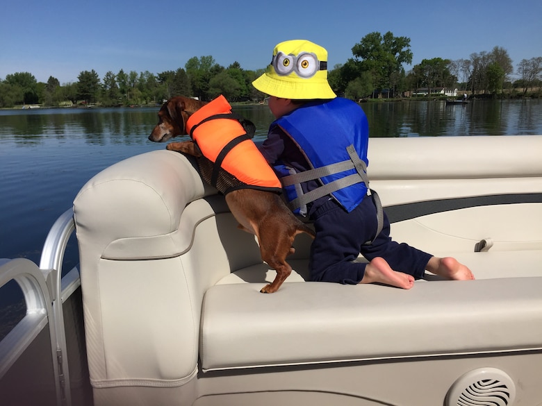 Child and dog, both wearing PFDs, on a pontoon, look over the water.