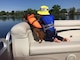 Child and dog, both wearing PFDs, on a pontoon, look over the water.