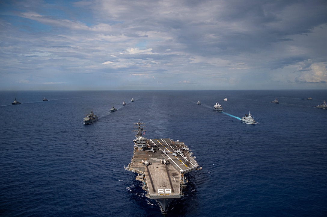 An aircraft carrier leads a formation of 13 other ships in the deep blue ocean, with the horizon in the background.