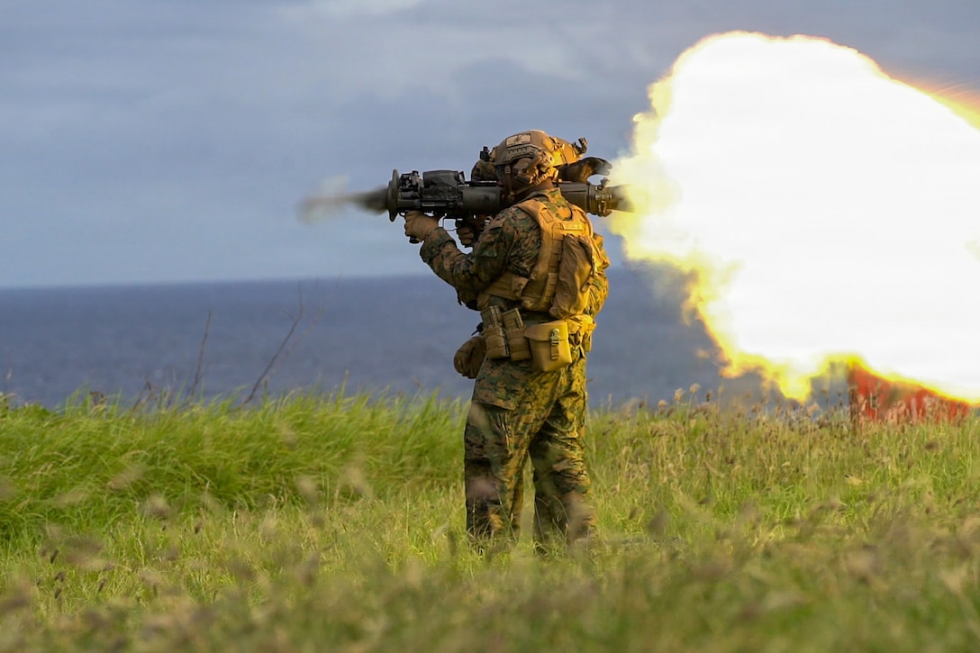 A fireball shoots out the back of a shoulder-fired weapon as a Marine fires it in a field.