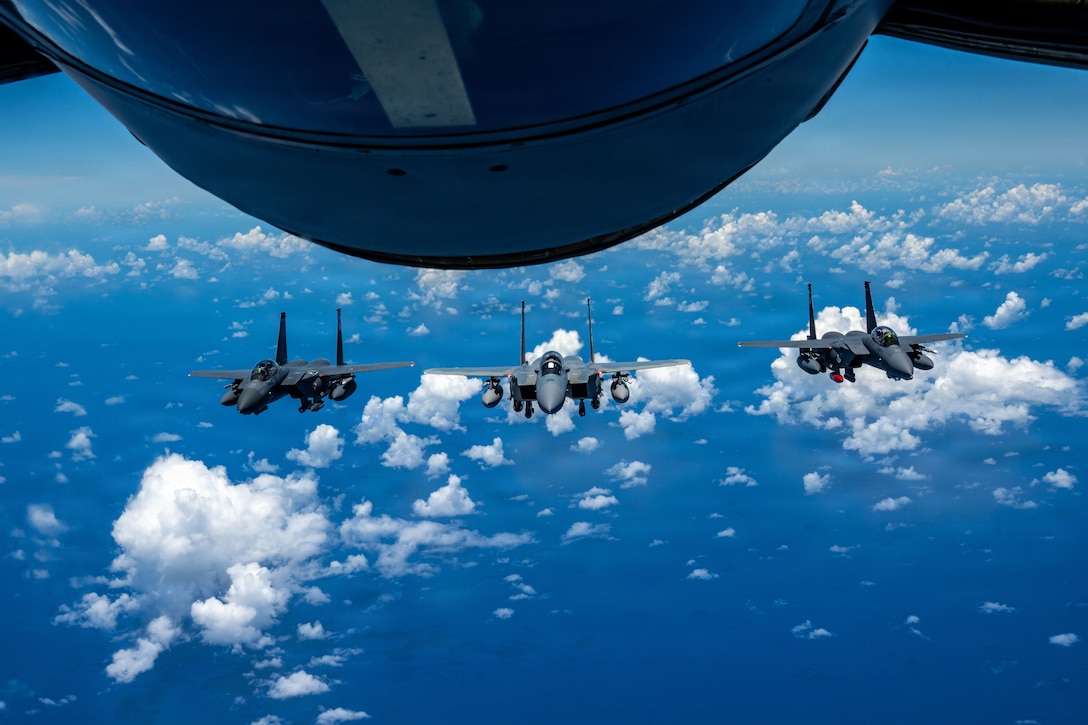 Three fighter jets fly in a row below and behind a refueling aircraft in the Pacific blue sky. Small bright white clouds appear as if painted on the sky.