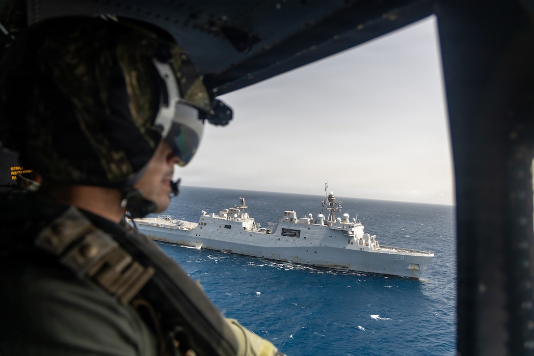 A man on a helicopter looks out an open door at a naval ship in the water.