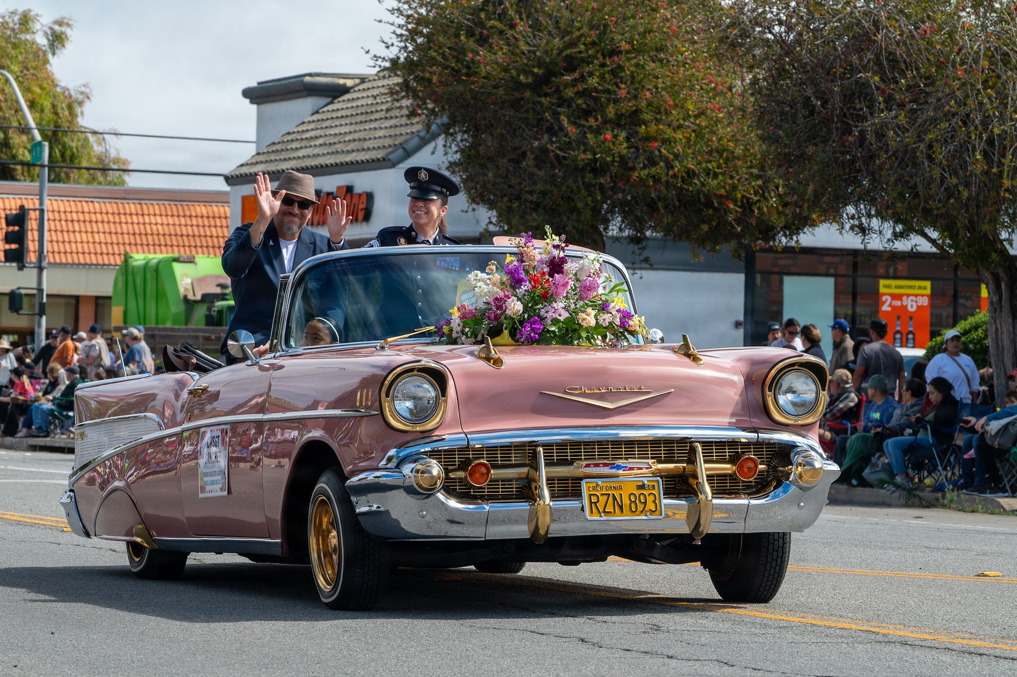 Two people sit in a car and wave at a crowd